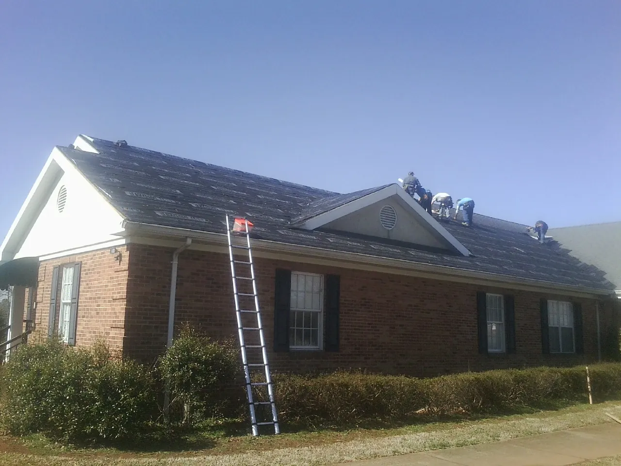 Crew tearing shingles off a brick ranch roof with a ladder set — Emergency Roof Repair work by our crew in Hanley Falls