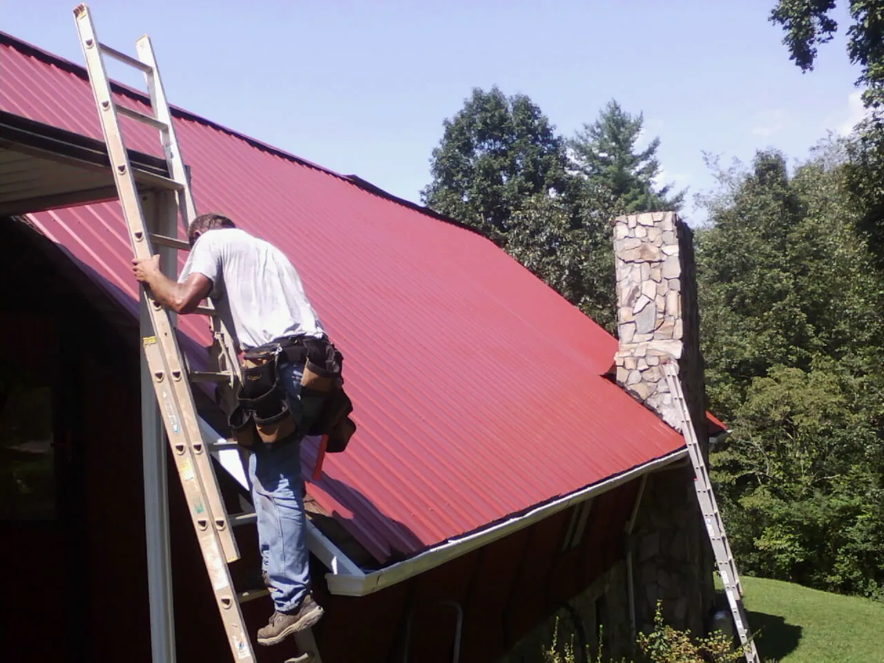 Roofer on a ladder beside a red metal roof and stone chimney — Soffit & Fascia Repair by Roofing Brothers in Hanley Falls